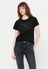 Svart bomull t-skjorte med rund hals og et teksturert, metallic grafisk design foran. Kombinert med mørk grå jeans.