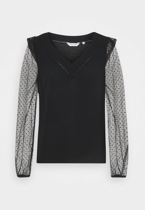 Haut noir avec un col en V et des manches grises transparentes à pois. Comprend des accents de tissu texturé au niveau du col et des manches fluides.