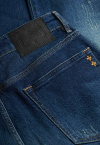 Jeans de mezclilla azul oscuro con costuras naranjas, un bolsillo trasero con dos pequeñas cruces naranjas, y una etiqueta de marca Superdry negra encima del bolsillo.