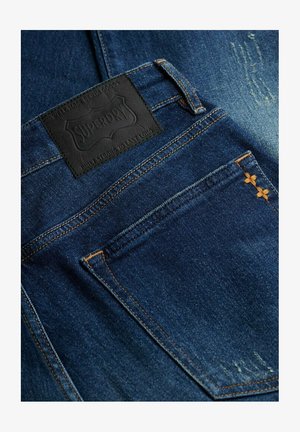 Donkerblauwe denim jeans met oranje stiksels, een achterzak met twee kleine oranje kruissteekjes en een zwart Superdry merklabel boven de zak.