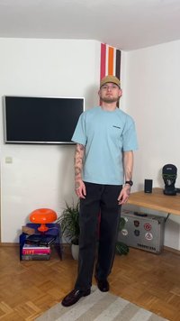 T-shirt azul claro oversized, calças pretas de corte reto e sapatos escuros. Tatuagens visíveis nos braços. Decoração e mobiliário minimalistas ao fundo.