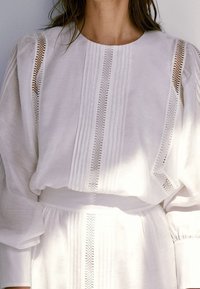 Blouse blanche en lin avec un design plissé et des accents en tissage ouvert. Elle présente un col rond et des manches volumineuses. Simple et texturée.
