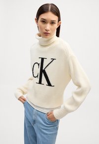Cremfarbener Rollkragenpullover mit auffälligem schwarzem "CK"-Logo, gerippter Textur, verkürztem Design und langen Ärmeln. Kombiniert mit hellblauen Jeans.