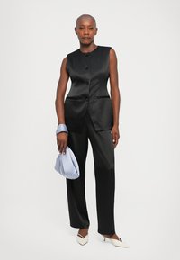 Sieviete ar noskūtu galvu valkā melnu bezpiedurkņu veste ar pogām, atbilstošas bikses, baltus zābakus un tur gaismas zilas rokas somiņas uz baltas fona.