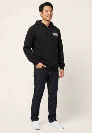 Mand smiler, iført sort hættetrøje, mørke jeans og hvide sneakers, står med den ene hånd i lommen mod en lys baggrund.