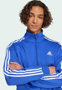 Blå zip-up träningsjacka med vita ränder längs armarna och en adidas-logga på bröstet. Tillverkad av ett mjukt och slätt tyg.