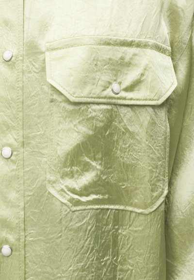 Φιστικί κατσαρό υφασμάτινο πουκάμισο με τσέπη με καπάκι ασφαλισμένη με κουμπί-πατητήρι και ορατές κούμπωμα κουμπιά μπροστά.