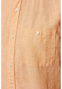 Camisa de lino naranja claro con un bolsillo en el pecho y botones blancos en la parte delantera, con una superficie ligeramente texturizada.
