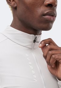 Gros plan d'une personne fermant une veste gris clair à col montant avec le nom de la marque « Isadore » visible sur la poitrine.