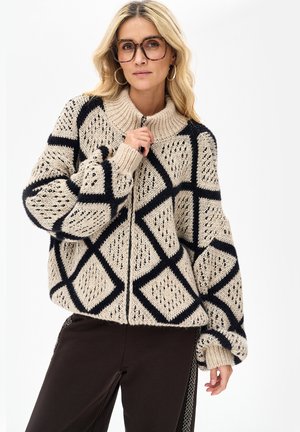 Kobieta w dużym beżowym swetrze z dzianiny z czarnym wzorem w romby, dużymi okularami i kolczykami w kształcie koła, pozująca na tle gładkiej białej ściany.