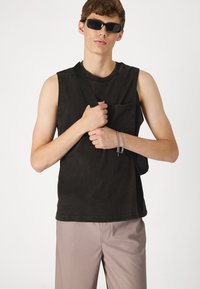 Débardeur noir sans manches avec une poche avant, porté avec un pantalon léger à rayures. Le modèle tient un sac à épaule noir et porte des lunettes de soleil sombres.