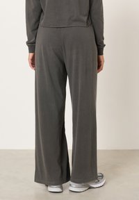 Asmuo dėvi laisvus tamsiai pilkus kelnes ir pilkus sportbačius, stovintis ant šviesiai pilkos grindų prie paprastos smėlio spalvos sienos.