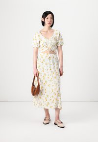 Bloemrijke crop top met strikdetail en pofmouwen, gecombineerd met een lange rok. Witte stof met gele bloemen; bruin handtas en lichte schoenen.