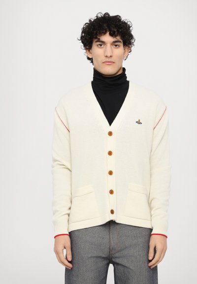 Cremefarvet cardigan med v-hals og træknapper. Har røde detaljer på ærmerne, to frontlommer og ribbede manchetter. Bæres over en sort turtleneck.