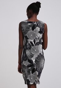 Kvinna med mörk hud och hår i en knut, klädd i en ärmlös svart, vit och grå klänning med abstrakta cirkulära och bladmönster, sedd bakifrån.