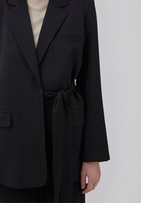 Sort blazer med bælte, der har et struktureret design, lange ærmer, to forlommer og en enkelt brystlomme med en glat tekstur.
