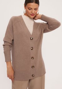 Jasnobrązowy kardigan dzianinowy z dekoltem w serek, prążkowanymi mankietami i guzikami z przodu. Charakteryzuje się miękką teksturą i luźnym krojem.