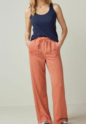 Frau trägt ärmelloses marineblaues Tanktop und korallenfarbene, weite Hosen mit elastischem Bund, steht mit den Händen in den Taschen.