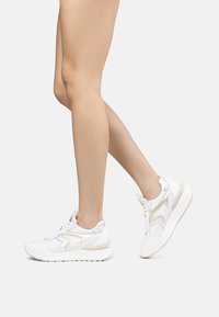 Sneakers atletici bianchi con un mix di materiali lisci e testurizzati, suola imbottita e accenti metallico oro in un design contemporaneo.