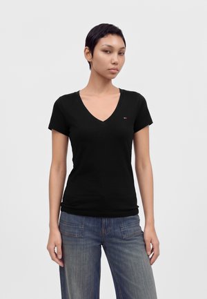 Jeune femme aux cheveux courts et foncés portant un t-shirt noir à col en V et un jean bleu, debout devant un fond clair uni.