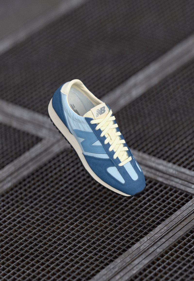 Blue athletic shoe with a smooth upper, suede accents, and cream laces. Features a lightweight outsole and layered design elements.