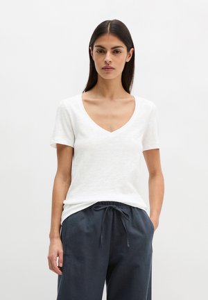 Frau trägt ein weißes T-Shirt mit V-Ausschnitt und kurzen Ärmeln sowie dunkelblaue Hose mit Kordelzug, steht mit einer Hand in der Tasche vor schlichtem Hintergrund.