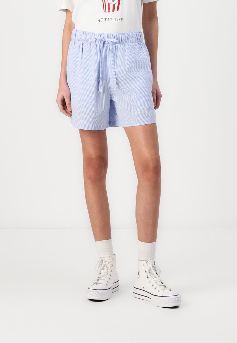Pantaloni corti a righe blu e bianche con vita elasticizzata e stringa, caratterizzati da una vestibilità ampia e un tessuto leggero, abbinati a sneaker bianche.