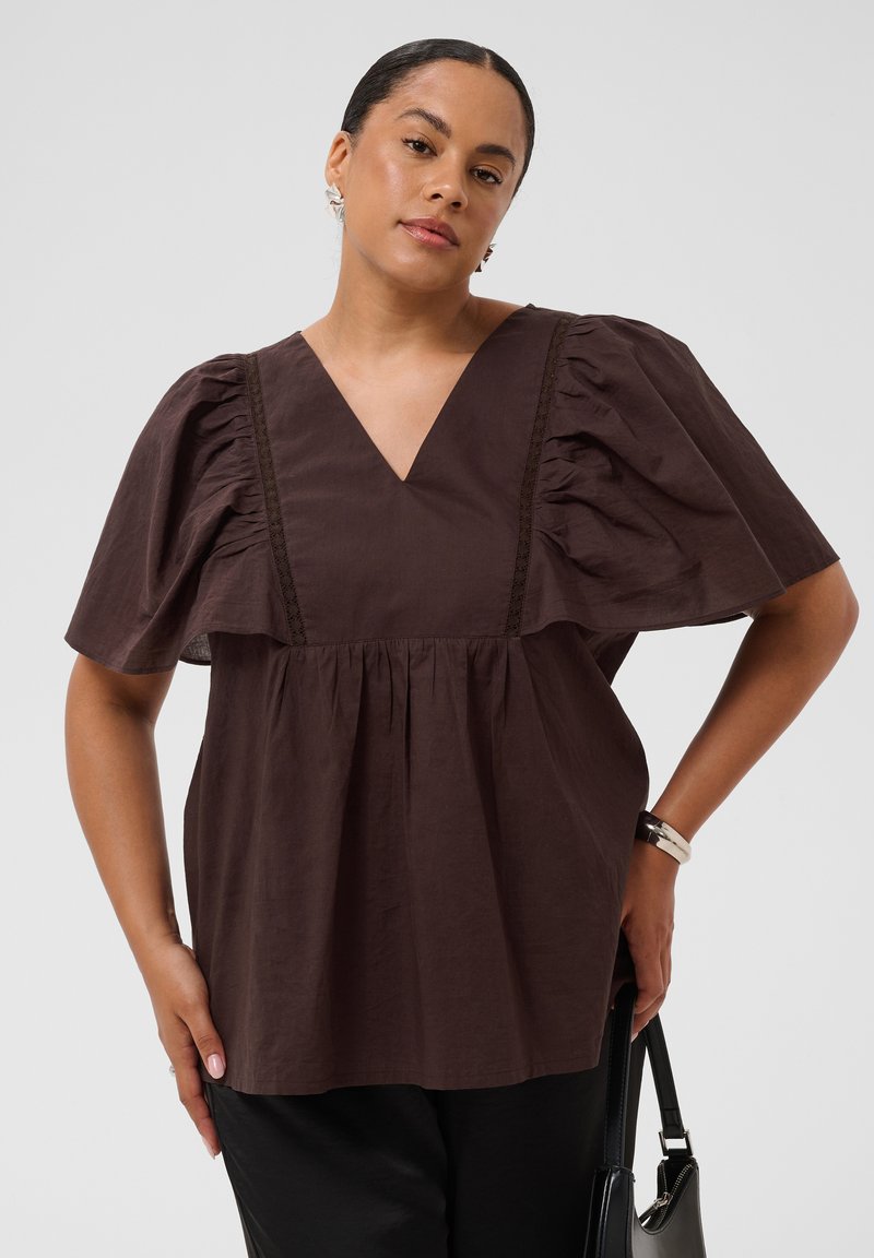 Femme portant un chemisier marron foncé à col en V avec des manches bouffantes, un pantalon noir, des boucles d'oreilles et un bracelet argentés, tenant un sac à main noir.