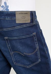 Tummansiniset farkut, joissa on kulunut pinta, varustettu "JACK & JONES" -nimilapulla ja koristeommelluilla takataskuilla.