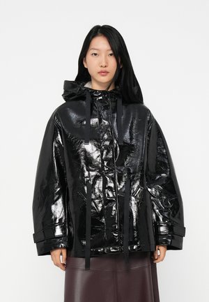 Donna con lunghi capelli neri che indossa un giubbotto nero lucido e oversize con cappuccio e una gonna di pelle marrone scuro, rivolta in avanti.