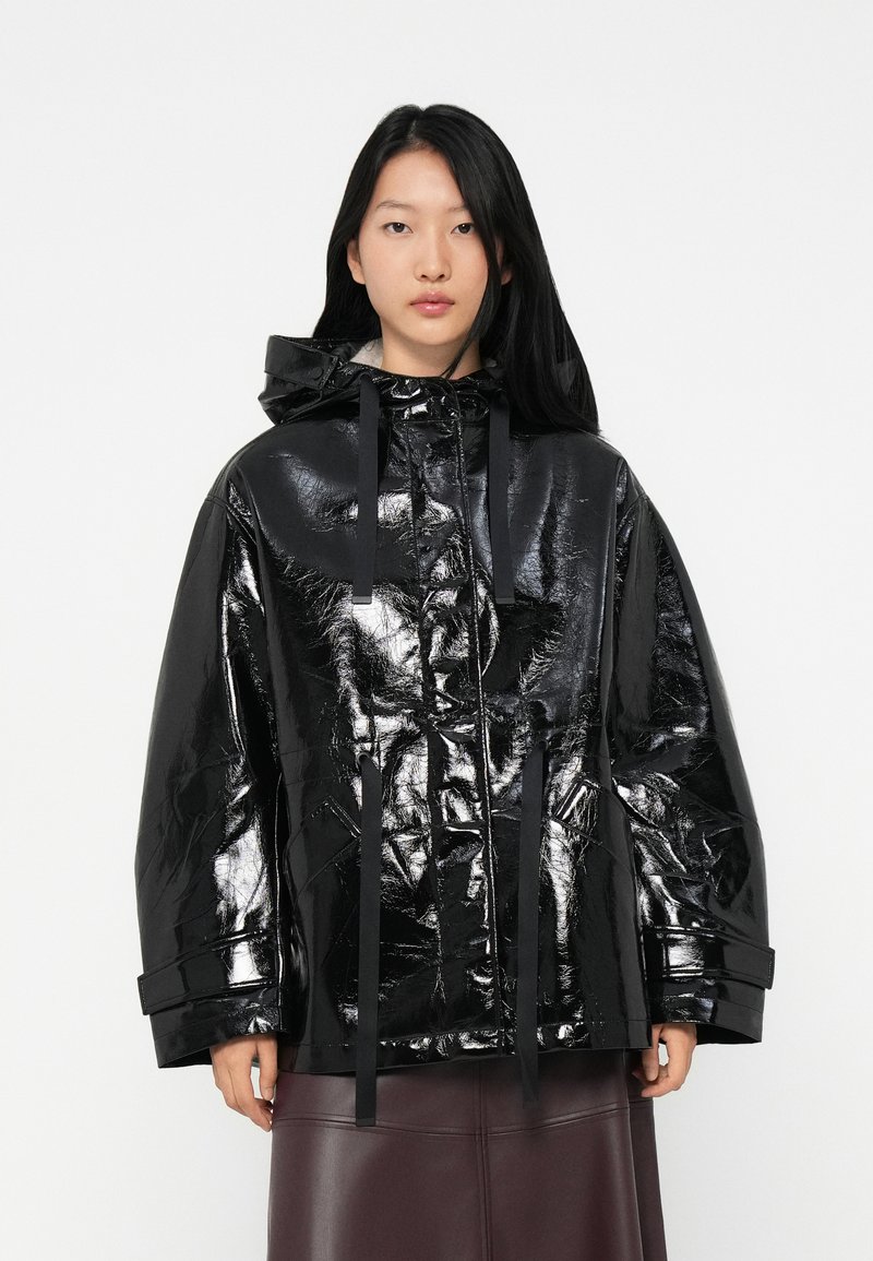 Femme avec de longs cheveux noirs portant une veste à capuche brillante et surdimensionnée noire et une jupe en cuir brun foncé, faisant face devant.