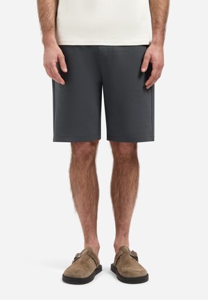 Man draagt donkergrijze knielange shorts, beige instapschoenen en een wit shirt tegen een effen lichte achtergrond.