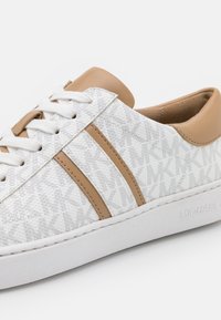 Baskets basses blanches et beige avec motif monogramme MK, détails en cuir beige, lacets blancs et semelle épaisse blanche embossée du nom de la marque.