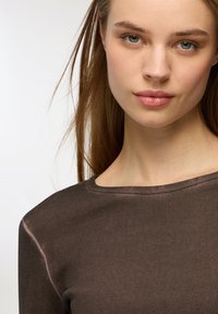Długi rękaw, prążkowany top w ciemnym brązie. Posiada szeroką, okrągłą dekolt i miękką teksturę. Włosy swobodnie opadają, otaczając twarz.