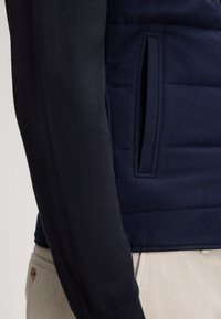 Granatowy sweter z pikowanym wzorem, z gładkim rękawem dzianym. Posiada boczną kieszeń oraz ściągacze przy mankietach dla komfortu i dopasowania.