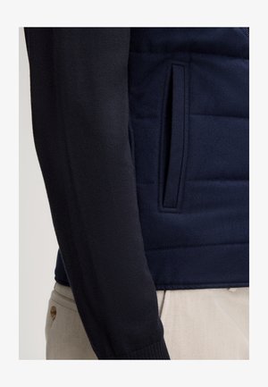 Granatowy sweter z pikowanym wzorem, z gładkim rękawem dzianym. Posiada boczną kieszeń oraz ściągacze przy mankietach dla komfortu i dopasowania.