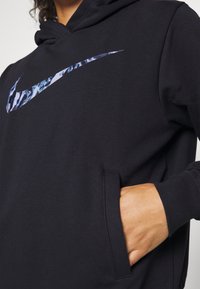 Osoba oblečená v černé mikině s kapucí, na které je modře vzorované logo Nike swoosh, ruka v přední kapse, ukazující texturu látky a detaily šití.