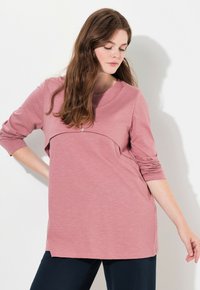 Top largo e morbido di colore rosa chiaro, caratterizzato da un yoke curvo con dettaglio bottonato e fessure laterali. Design semplice e sobrio.