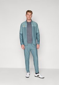 En teal idrettsdrakt laget av glatt stoff, med hvite aksentstriper, glidelåsjakke og slanke bukser. Båret med hvite joggesko.