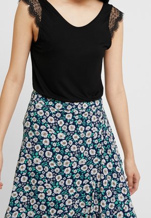 Frau trägt ein ärmelloses schwarzes Oberteil mit Spitzen an den Schultern und einen marineblauen Rock mit weißem Blumenmuster.