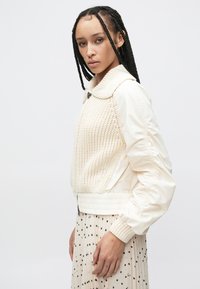 Biała bomberka z dzianinową fakturą na górnej połowie, pufiastymi rękawami, ściągaczami na mankietach i szerokim kołnierzem. Nosi się ją na spódnicę w groszki.