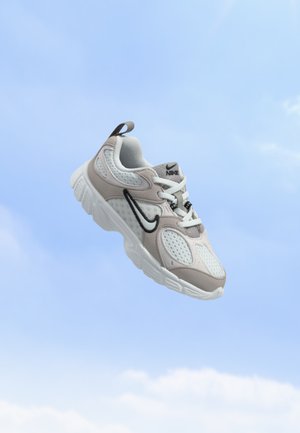 Baskets Nike blanches et beiges avec un logo Swoosh noir flottant sur un fond de ciel bleu.