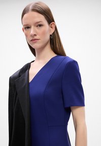 Blå kjole med korte ermer og V-hals, kombinert med en svart blazer på den ene skulderen. Glatt tekstur og strukturerte designdetaljer er synlige.