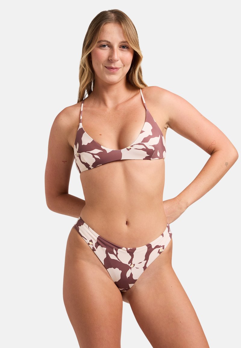 Femme aux cheveux blonds portant un bikini marron et crème à fleurs, debout avec une main sur la hanche, devant un fond clair uni.