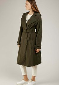 Manteau trench vert olive avec une taille ceinturée, des boutons double boutonnage et un grand col. Porté sur une chemise blanche et un pantalon clair.