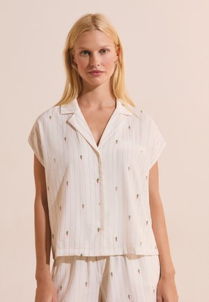 Blonde vrouw die een wit overhemd met korte mouwen en knopen draagt, en bijpassende korte broek met dunne rode strepen en kleine radijsprints.
