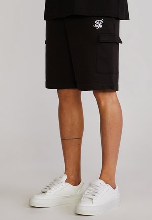 Person trägt schwarze Cargo-Shorts mit Logo auf der Tasche und weiße Sneakers, steht vor neutralem Hintergrund.