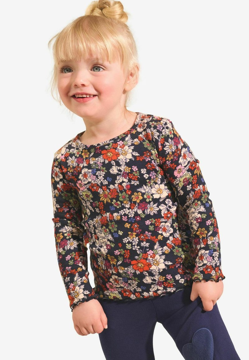 Langarmshirt in Marineblau mit einem mehrfarbigen Blumenmuster. Gerippte Textur und Rüschensaum. Kombiniert mit dunkelblauen Leggings mit einem Herz-Patch.
