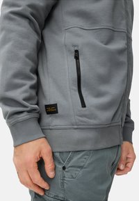 Grijze sweatvest met ritssluiting, zijzak, geribbelde manchetten en zoom; voorzien van een stoffen label met zwarte en oranje tekst.