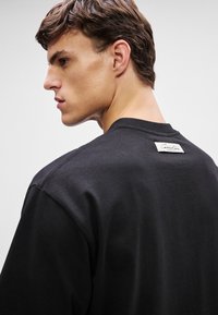 Jovem com cabelo castanho curto, vestindo um moletom preto da Calvin Klein, de perfil para a esquerda com uma expressão neutra num fundo branco.